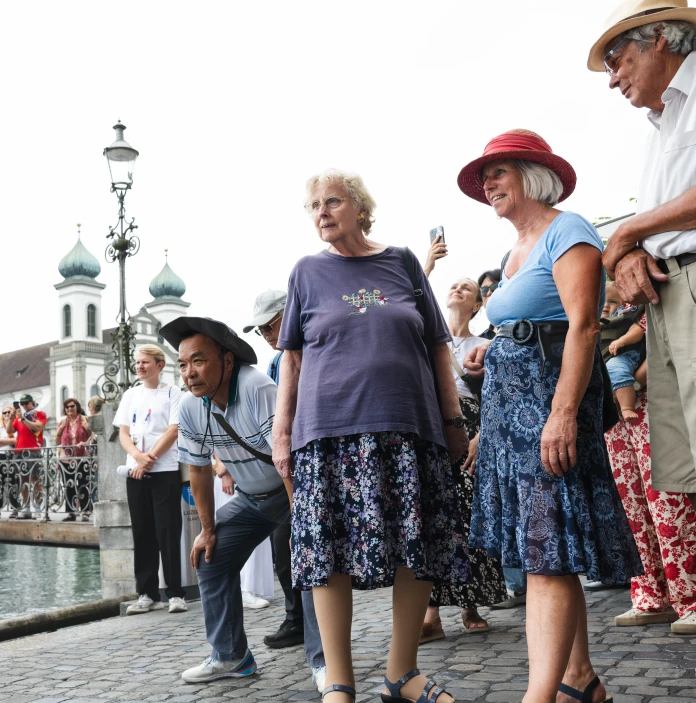 A group of tourists gather beside a riverside bridge, with historic buildings in the background.