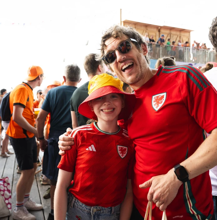 A smiling adult and child wearing red Wales football shirts pose together among a crowd of fans.