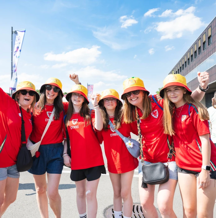 A group of smiling Wales football fans in red shirts and colourful hats pose together outside a stadium.