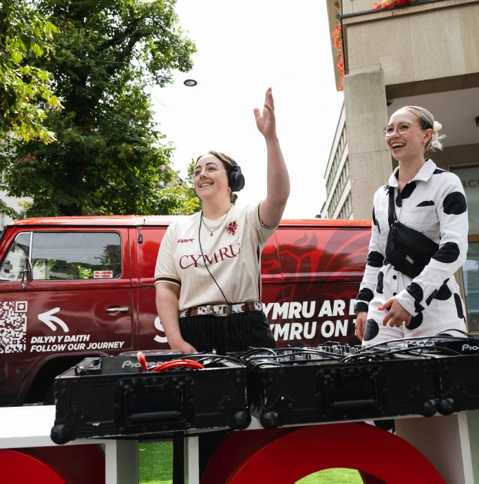 Two women smile behind DJ equipment at an outdoor Wales football event.