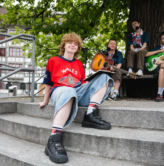 A group of musicians in Wales shirts pose with guitars beneath a large tree.
