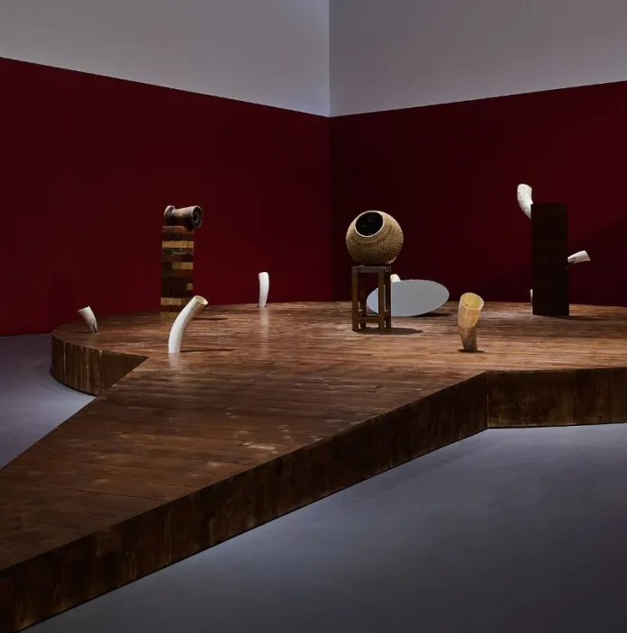The installation showcases a sculptural arrangement on a wooden platform, featuring abstract, geometric forms set against a contrasting red and white wall backdrop