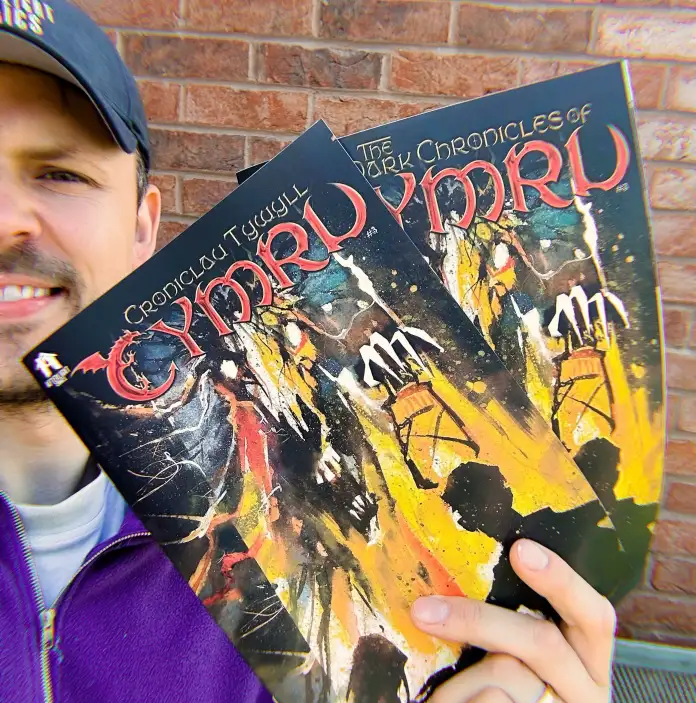 A person holding two copies of The Dark Chronicles of Cymru comic book in front of a brick wall.