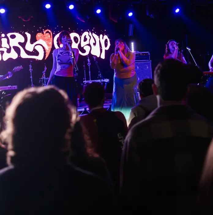 A group of performers singing on stage with an audience watching. Colorful lighting highlights the performance.