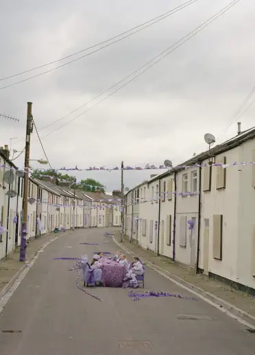 A quiet residential street with rows of boarded-up houses stretches into the distance under a cloudy sky. In the middle of the road, a small group sits around a table decorated with purple ribbons and balloons.