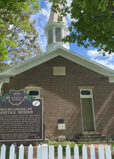 The outside of a historic brick building is surrounded by trees and a white fence. A sign stands in front of the building near the entrance. 