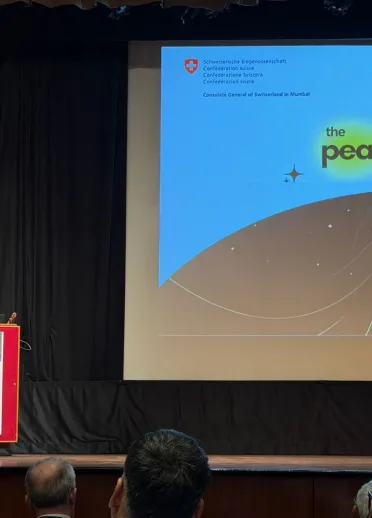 Two speakers at a lectern on stage during an event for The Peace Particle, with the CERN documentary film’s title displayed on a large screen behind them.