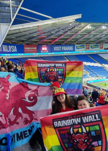 A group of Welsh football fans holding up rainbow banners in a football stadium
