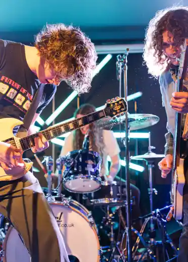 Two musicians playing electric guitars on stage with bright lights behind them. A drummer is visible in the background.