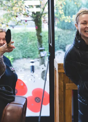 Lauren Price MBE laughs while visiting a local barbershop, as a barber cuts a customer's hair in the foreground.