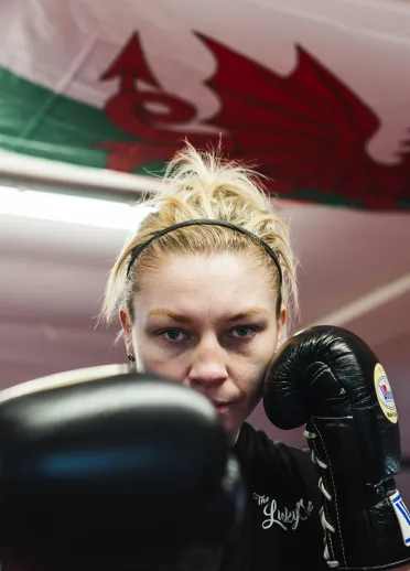 Lauren Price MBE in a boxing gym, wearing black training gear with her gloves raised, extending one fist towards the camera during a training session.