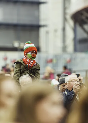 Young Welsh rugby fan wearing a red, white and green hat and scarf, with a Welsh dragon painted on their face, sitting on someone's shoulders in a crowd outside the Principality Stadium. 