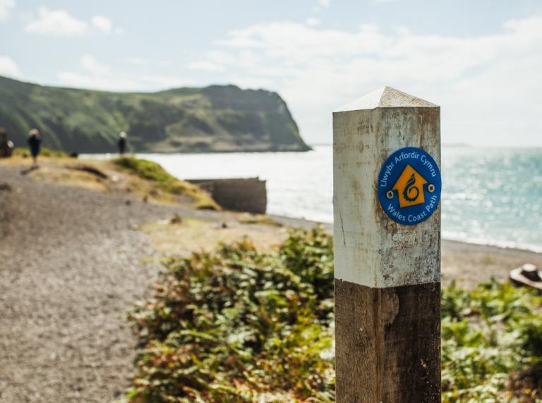 A close-up sign showing the direction of the Wales Coast Path in front of a beachside trail 