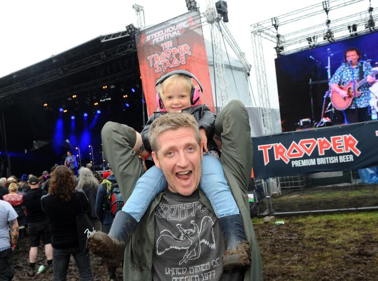A man standing next to the main stage with a child on his shoulders wearing ear protectors.
