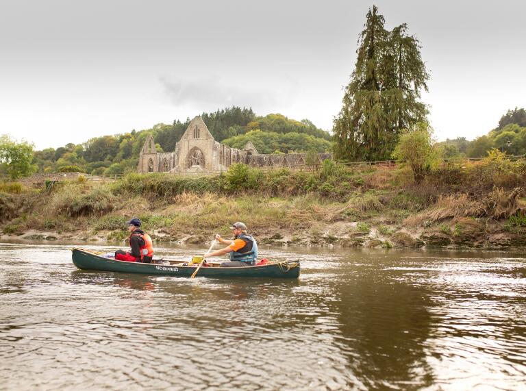 Two people canoeing on the river past an abbey ruins.