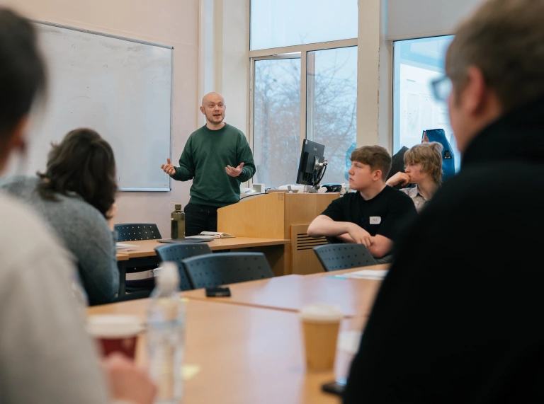 A person stands at the front of a classroom speaking to a room full of young people. 