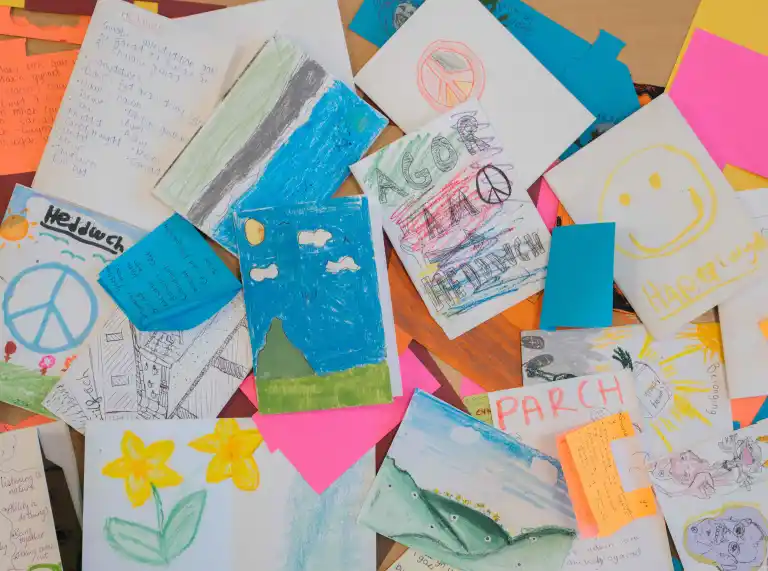 A collection of colourful drawings and handwritten notes spread across a table. The artwork includes peace symbols, landscapes, flowers, and positive messages. 