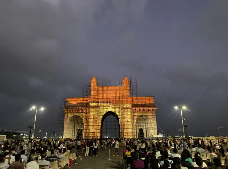 The Gateway of India illuminated at night, surrounded by scaffolding, with a large crowd gathered in front beneath a dark, cloudy sky.
