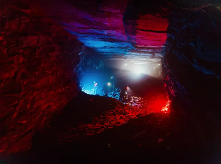 Revellers making their way through a vast underground slate cavern illuminated by red and blue lights. 