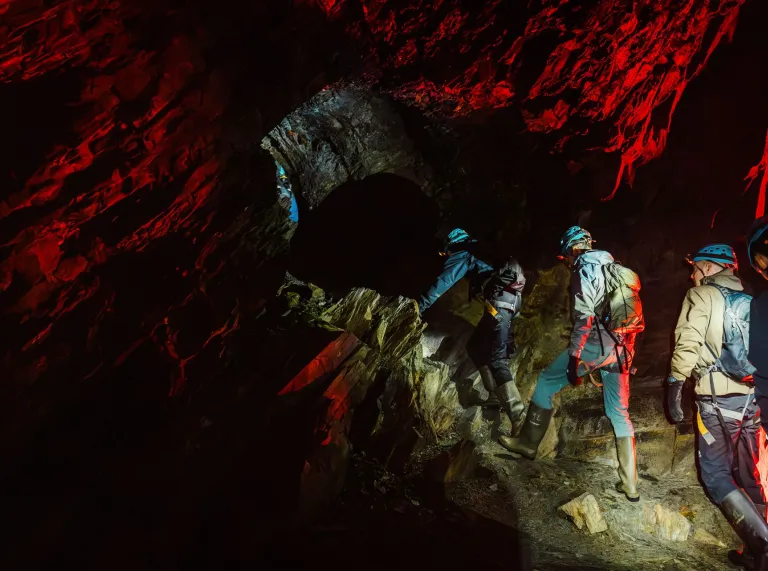 Helmeted revellers climbing through a rocky tunnel illuminated by red lights at Cwmorthin Slate Mine. 