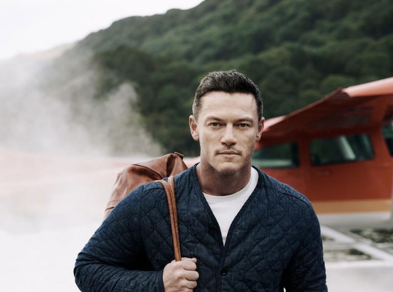 Luke Evans walking out of small aircraft with smoke behind him