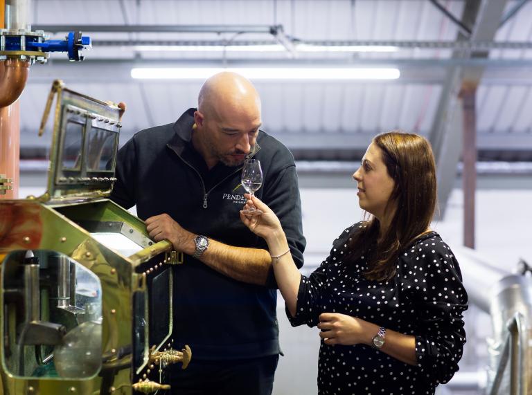 man leaning on machinery smelling whisky as woman holds glass for him 