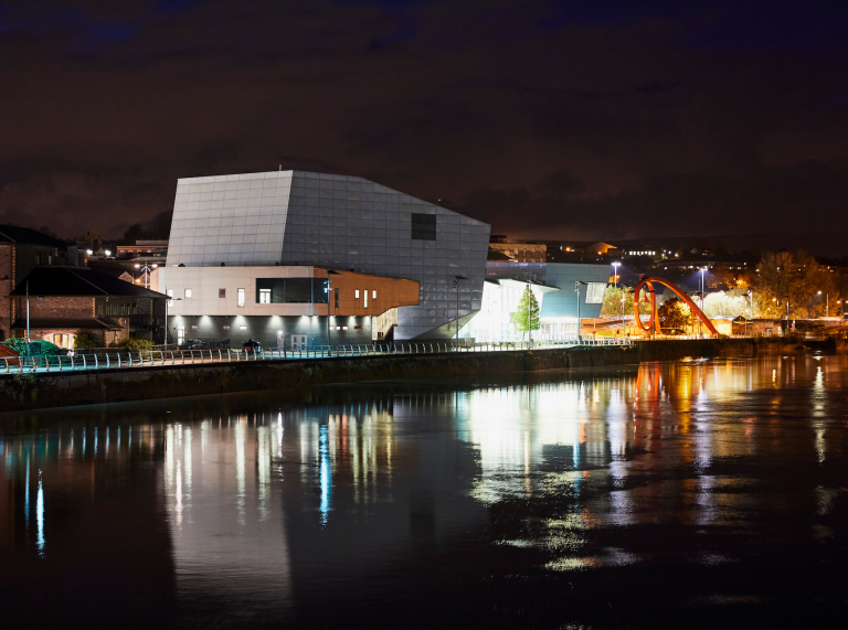 Foto nocturna de la orilla del río, con luces del edificio que se refleja en el río