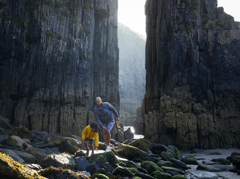 father and son scrambling over rocks with cliffs in background.