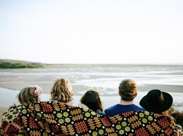 backs of heads of five people wrapped in colourful blanket with sea in background.