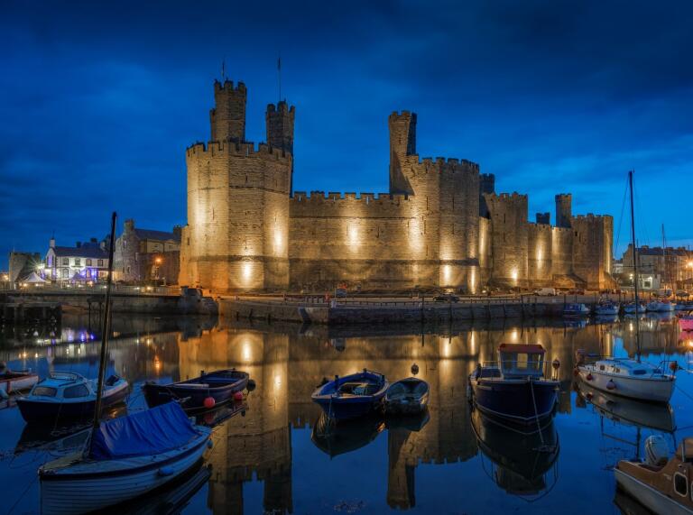 A grand old castle with a moat and boats in the foreground, lit up against a night sky