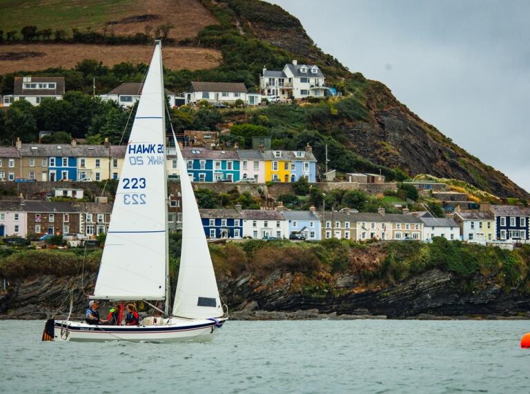 A sailing boat on the sea with a coastline of colourful houses up a green hill in the background
