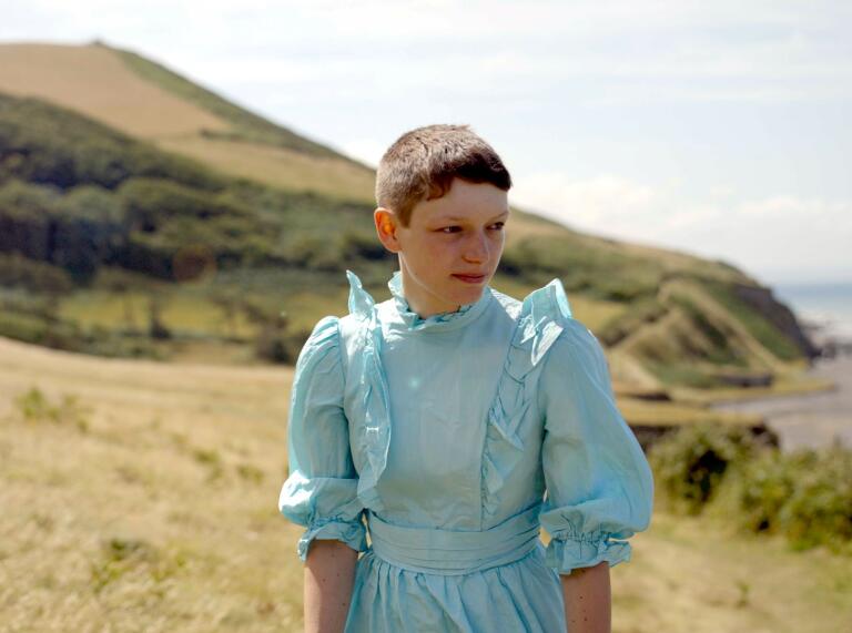 A portrait of a person in a blue dress standing in a green mountainous landscape