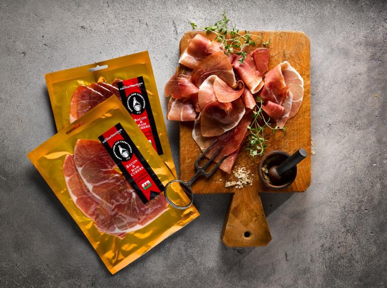 Two packets of ham and a wooden board displaying carved ham.