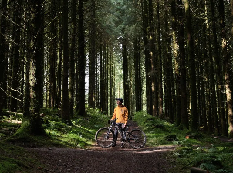 Una persona con una chaqueta amarilla y equipo de ciclismo de pie con una bicicleta de montaña en un camino rodeado de árboles altos en un hermoso entorno forestal