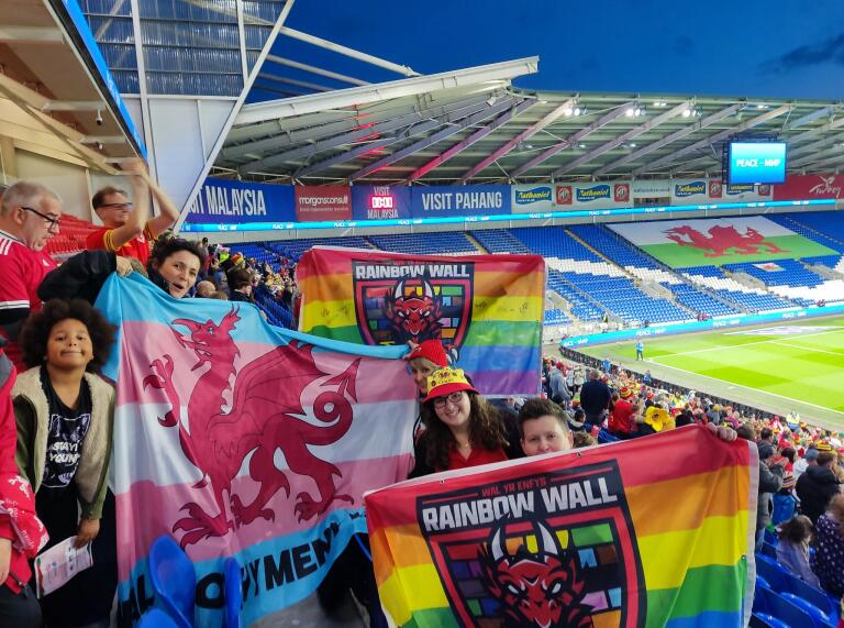 A group of Welsh football fans holding up rainbow banners in a football stadium