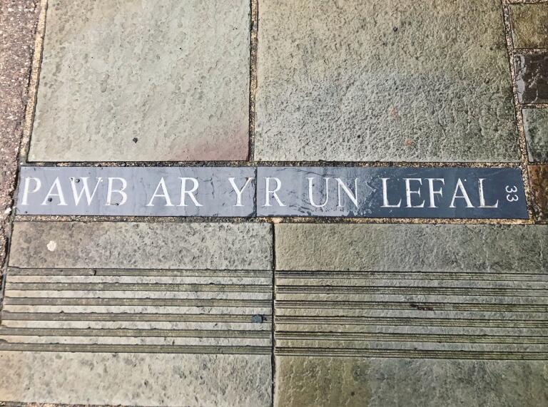 Pawb ar yr un Lefel', written in a piece of slate embedded into a slabbed pavement.