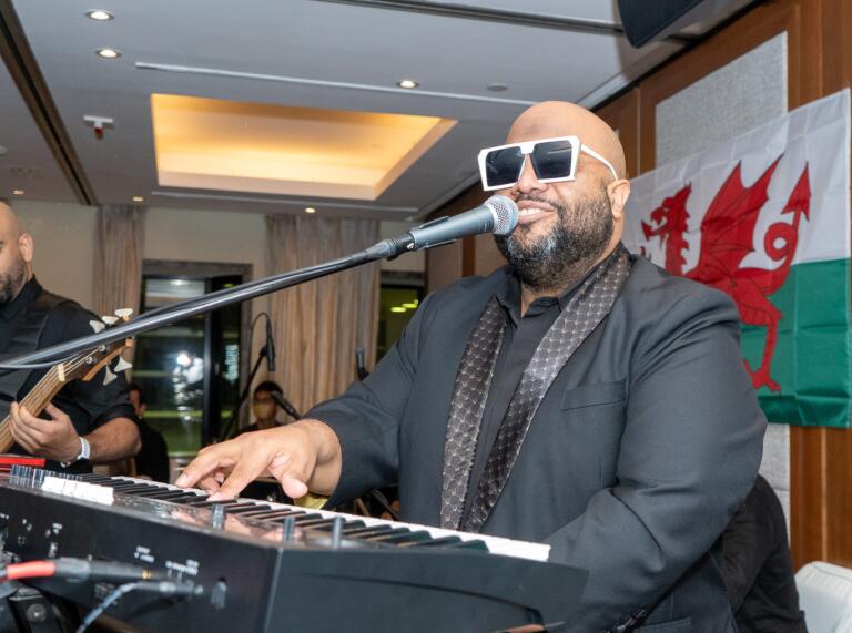 A man wearing sunglasses and a smart suit sitting playing a keyboard, with a Welsh flag in the background