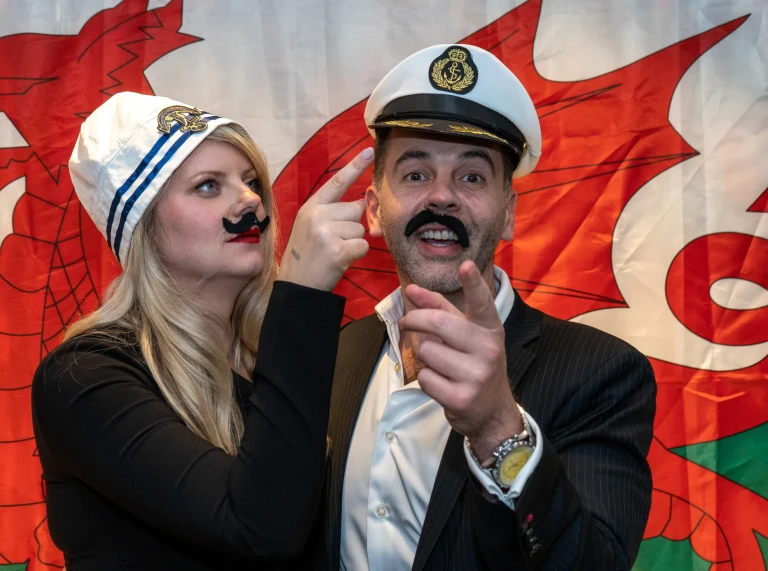 Deux personnes portant des chapeaux de marin et de fausses moustaches se tenant devant un drapeau gallois