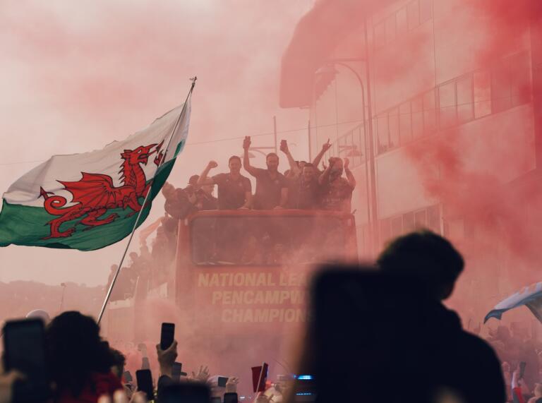 A tour bus with a sports team driving past a stadium, through throngs of fans waving flags and letting off flares