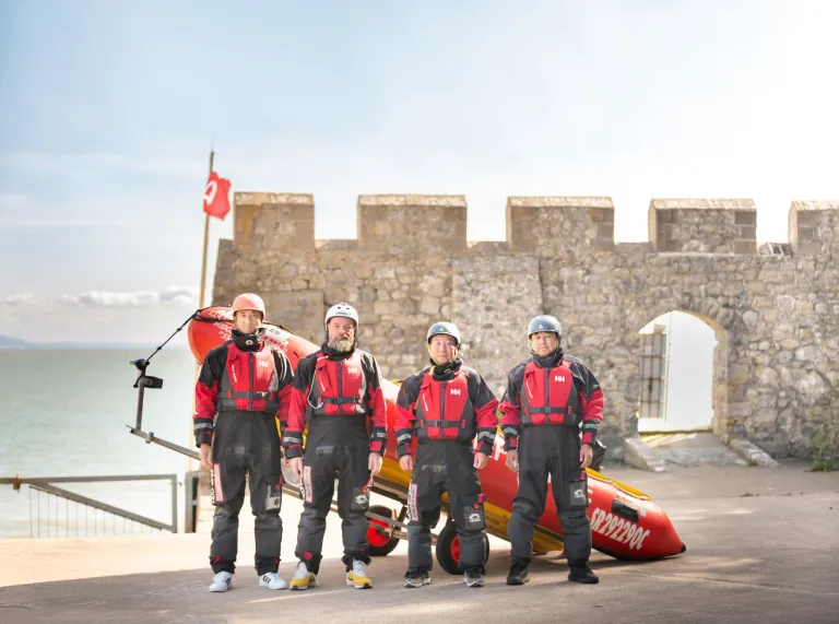 Four men dressed in sea rescue clothing stand in front of a stationary lifeboat on land in front of a castle wall