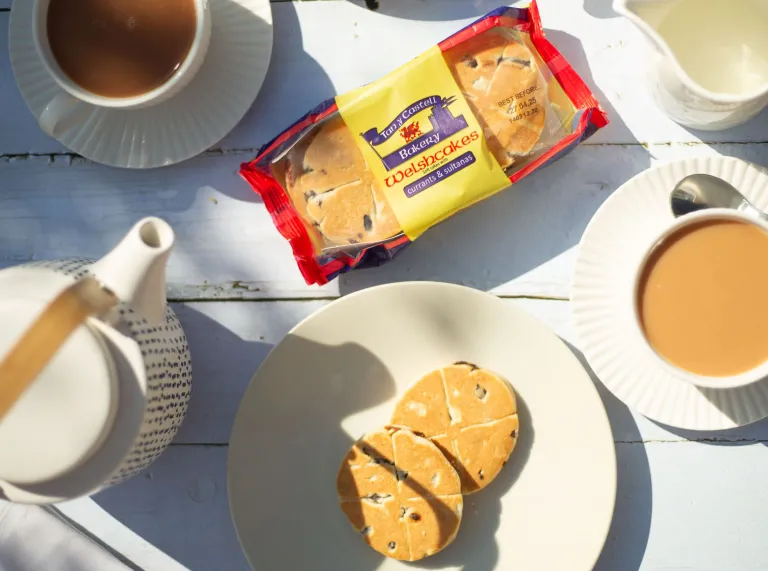 A packet of Tan y Castell Welsh cakes next to a cup of tea and some loose Welsh cakes on a saucer