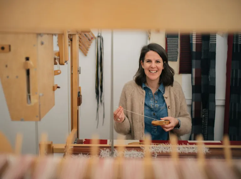 La artista textil galesa Llío James sentada en su telar manual, sonriendo a la cámara.