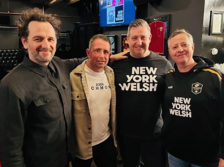 Un groupe de quatre hommes debout dans un bar, souriant à l'appareil photo, deux d’entre eux portent des maillots des New York Welsh et un autre porte un haut de style rugby.