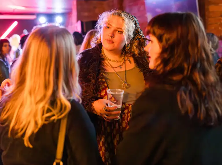 A small group of people chatting together in a dimly lit social setting. One person holds a drink while talking.