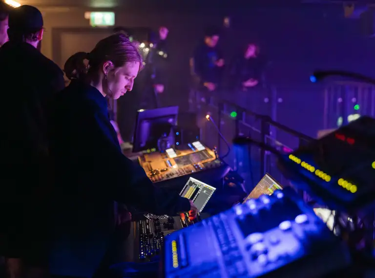 A person operating sound or lighting equipment in a dark venue. Screens and controls glow in front of them.