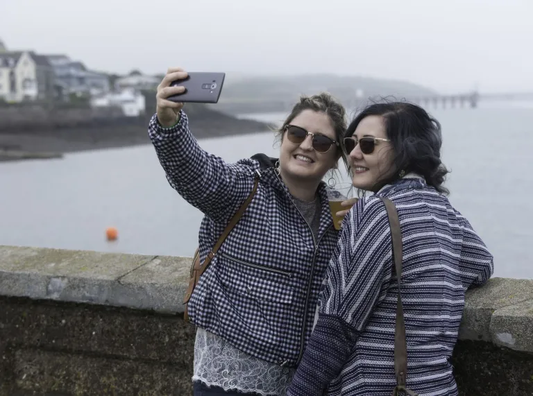 Two women wearing sunglasses taking a selfie beside a waterfront, with coastal buildings and a pier in the background. 