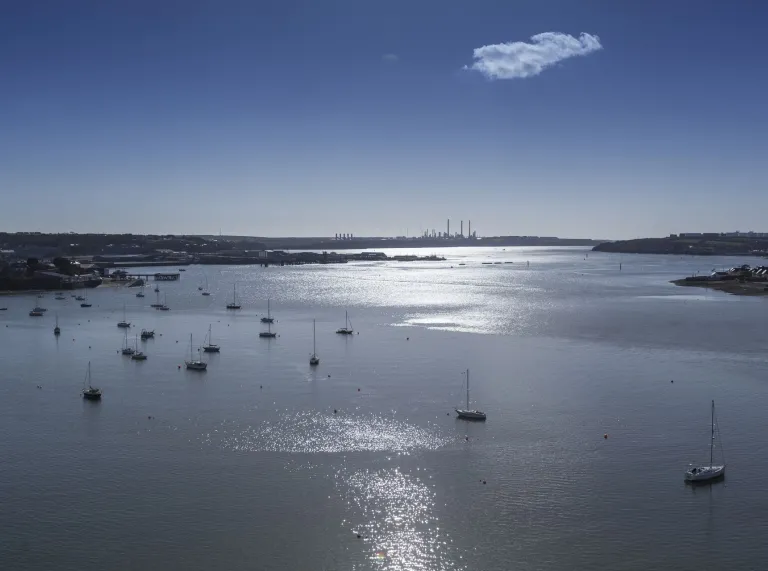 Sailing boats moored on the sunlit waters of Milford Haven in Pembrokeshire, West Wales, with the coastline and industrial skyline in the distance. 