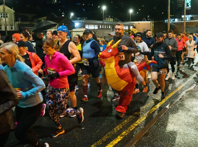 Runners taking part in the Nos Galan road race at night, including a participant wearing a colourful inflatable dragon costume. 