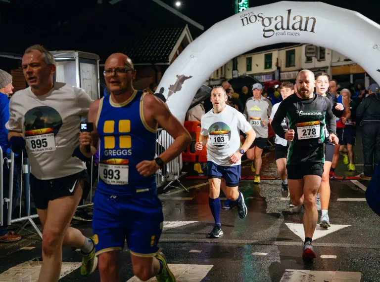 Runners passing beneath the illuminated Nos Galan Road Races arch during a night-time race on wet streets in Wales. 