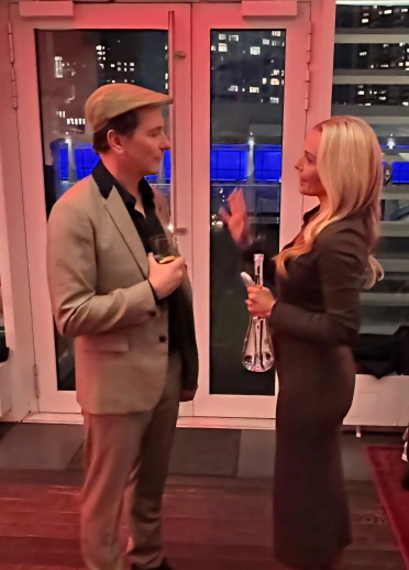 A man and a woman chat inside a dimly lit venue with city lights visible through the windows behind them.